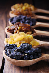 Black, brown and yellow raisins in spoons on a dark wooden background.