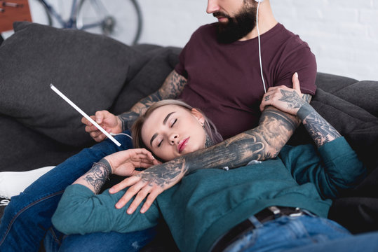cropped image of tattooed girlfriend sleeping and boyfriend listening music with tablet