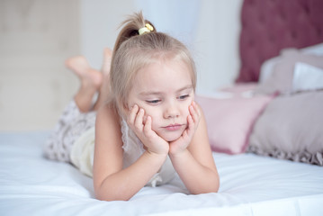 Sulky grumpy attractive little blond girl lying on a bed looking up into the air with a happy expression as she watches something.