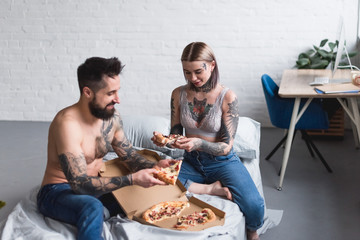 tattooed couple eating pizza on bed