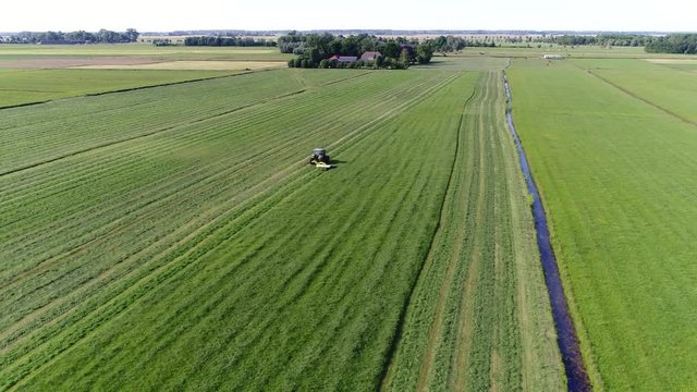 Aerial footage farmer cutting grass meadow and typical farm in background flight over the grass field showing farmer in tractor cutting the fresh green grass moving over the agricultural machinery 4k