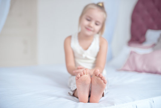 Little blonde girl sitting on the bed with focus on the feet indoors