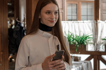Fototapeta premium a young girl with straight long hair and brown eyes in the restaurant holds the phone in her hands and thinks what message to write, smiles and looks aside