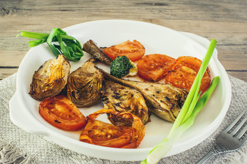 half artichoke, grilled and served with slices of ripe red tomatoes, Brussels sprouts and green onions on a white plate on a wooden background in rustic style.