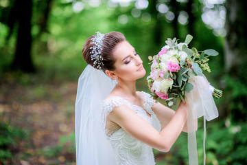 Naklejka premium Gorgeous bride holds pink wedding bouquet before her face posing in a green forest
