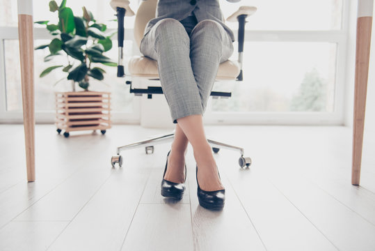 Cropped close up photo of healthy beautiful elegant woman's legs wearing high-heeled shiny black shoes, the woman is sitting in office at the table on modern luxurious armchair