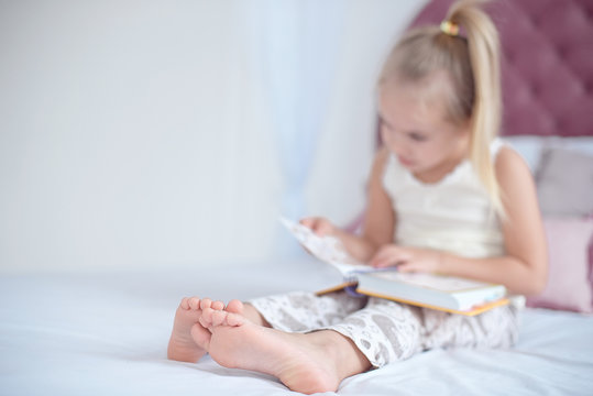 Little blonde girl sitting on the bed reading a book with focus on the feet indoors