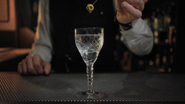 Cinemagraph of bartender holding up olive on sword stick over fancy and expensive alcoholic cocktail in vintage glass with dry martini dripping down from it