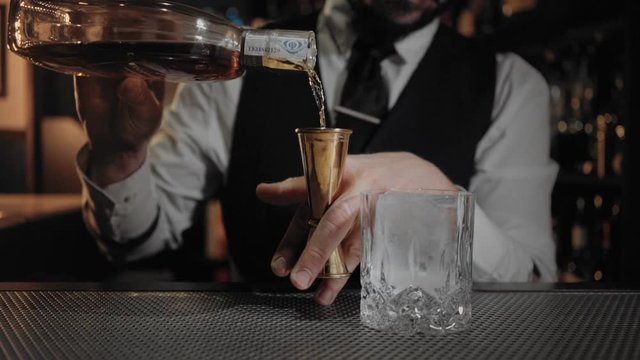 Professional and skilled artisan bartender at bar pours liquor or alcoholic drink into jigger and then in whiskey or scoth glass at expensive and invitation only establishment.