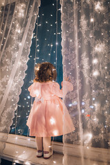 Cute child girl stay near the window and looking at the stars. Little kid stay at windowsill around garlands. Child like in fairytale. Curly child enchanted stars and sky.