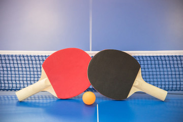 Orange ball for table tennis and two rackets of red and black color on a blue table with a grid