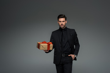 Stylish man in black suit holding gift box isolated on grey, international womens day concept