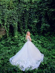 Blonde bride in rich wedding dress poses among greenery outside