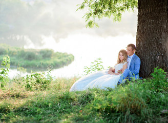 Bride and groom hold each other tender sitting under large old green tree