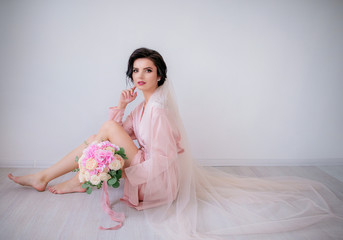 Fototapeta na wymiar Brunette bride in pink silk robe and long veil sits on the floor holding pink wedding bouquet