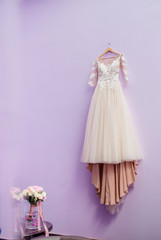 Wedding dress in color of dusty rose hangs on violet wall before the table with wedding bouquet
