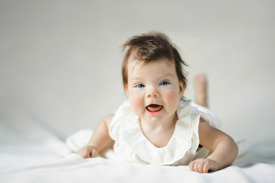 Gorgeous child with blue eyes in white dress lies on white bed in a bright room