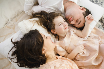 Mom and dad lie side by side with their little daughter on the soft white bed