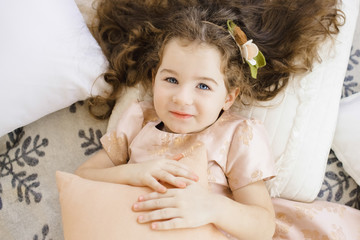 Curly little girl in peach dress lies on the soft bed