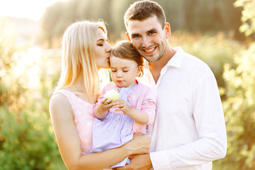 Happy young family spending time together in nature park. wife with husband and little baby. Mother and father kissing baby daughter.