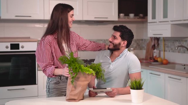 Cute couple in love having sweet talk in cozy kichen, bearded man, in light blue t-shirt, holding metallic tablet, beautiful long-haired woman, in stylish red shirt, standing nearby with shopping bag