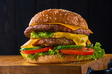 Closeup of home made beef burger with lettuce and tomatoes served on wooden cutting board. Dark background