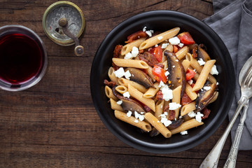 Italian wholemeal or wholegrain penne pasta with portobello mushroom, sun dried tomatoes, cherry tomatoes and feta cheese in a black bowl on dark rustic wooden table. Overhead view with copy space