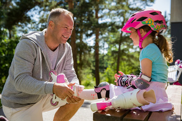 Father helping his daughter in helmet to wear protective pads for roller skates. Happy family...