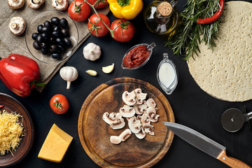 Cutting wooden board with traditional pizza preparation ingredients: cheese, tomatoes sauce, mushrooms, olive oil, pepper, spices. Black texture table background