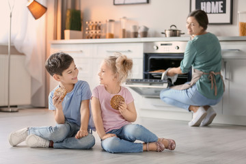 Little kids eating cookies near oven in kitchen while their mother cooking on background