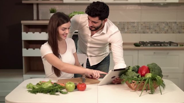 Great family evening, pretty dark-haired woman cutting tomato, handsome tall man showing pictures on tablet, indoor shot in fine white kitchen, concept of healthy eating and beautiful body