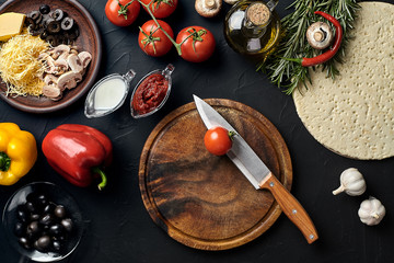 Cutting wooden board with traditional pizza preparation ingredients: cheese, tomatoes, sauce, olives, olive oil, pepper, spices. Black texture table background