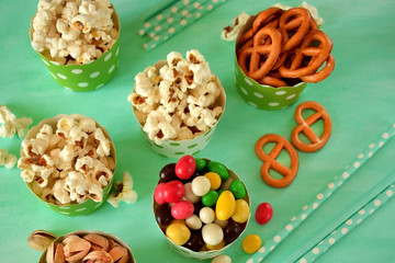 Popcorn, multicoloured drops, pretzels with salt and pistachio nuts in paper cups on green background. Snacks assortment