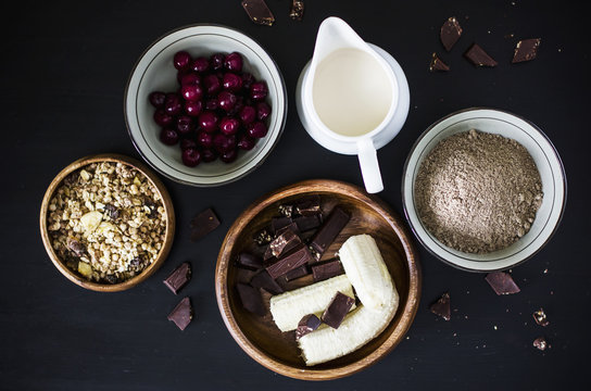 Muffins ingredients: milk, muesli, frozen cherry, flax flour, banana and chocolate on black background