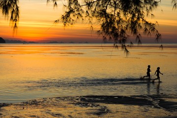 Couple of kids running freely on shallow water at sunset on the beach and tree branches falling from the top in the island of Koh Phangan, Thailand. Childhood innocence, happiness concept