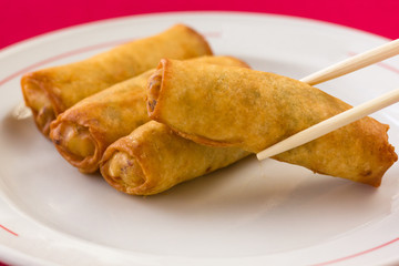 Chinese spring rolls on white plate and chopsticks grabbing another one over red tablecloth