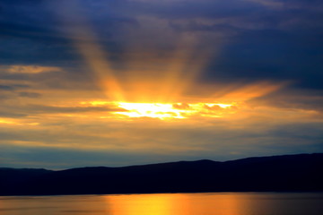 Dramatic sunset on a Bailkal lake. Bright yellow sun rays pass through the clouds and reflected in the water. Sunset landscape. Black silhouette of mountains in the background