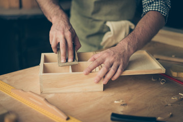 Close up cropped photo of handicraftsman's hands sanding the surface of handmade wooden box with abrasive paper