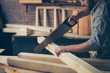 Cropped close up photo of concentrated serious professional hardworking qualified joiner sawing a wooden plank on his desktop with a handsaw