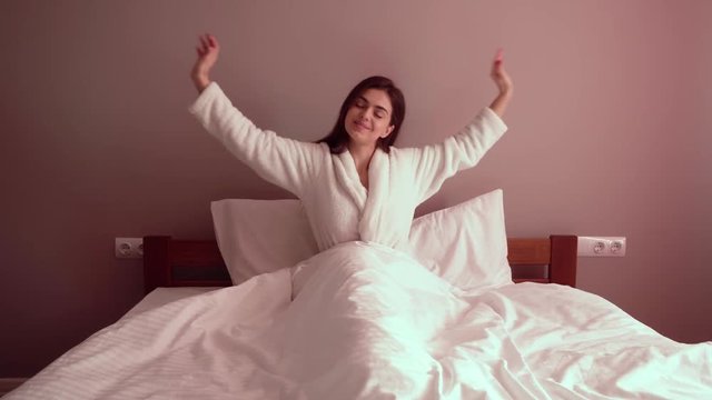 Pretty long-haired woman stretching in bed, wearing white soft bathrobe and elegant necklace, sitting in big bed under white blanket, waking up