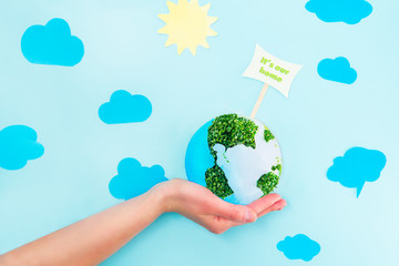 Female Hands holding Earth paper and green sprouts collage model with It's our home pointer on blue background with paper sun and clouds. Earth in your hands, Saving planet concept.