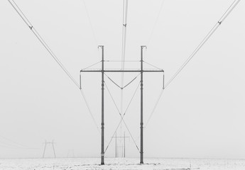 Hi-voltage electric power lines in a wintry foggy landscape, black and white photo.