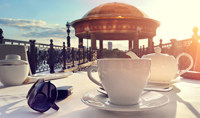 белая чашка на блюдце с белым чайником на белой скатерти на фоне золотой ротонды, а рядом лежат солнечные очки, ключ от машины и сахарница
