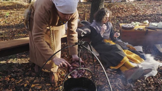A woman throws meat into a cauldron on an open fire in a Viking camp.