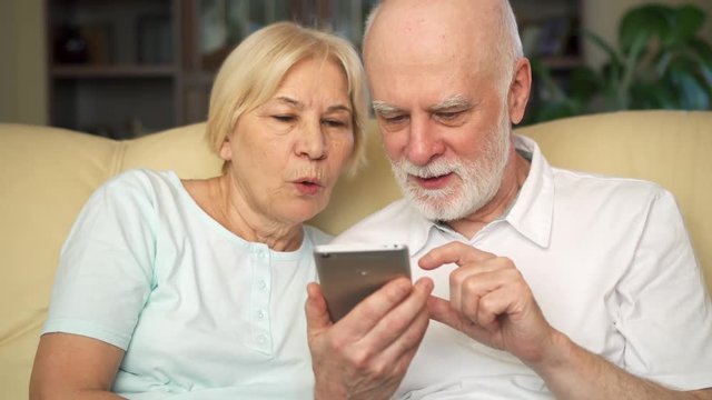 Cheerful good-looking senior couple sitting on couch at home. Modern pensioners using smartphone, browsing, reading news, looking at photos. Active modern life after retirement