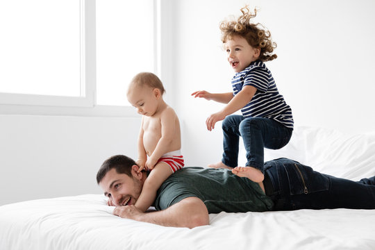 Dad lying on bed while son bounces on his back and baby straddles his neck