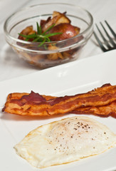 Top view of a freshly, fried egg, fresh ground pepper and two stripes of bacon on a round, white plate, with a clear glass bowl of roasted red potatoes,  rosemary and a fork 