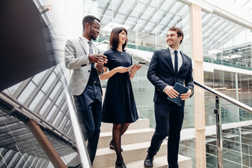 Three happy multiracial business people walking down on stairs together with digital tablet