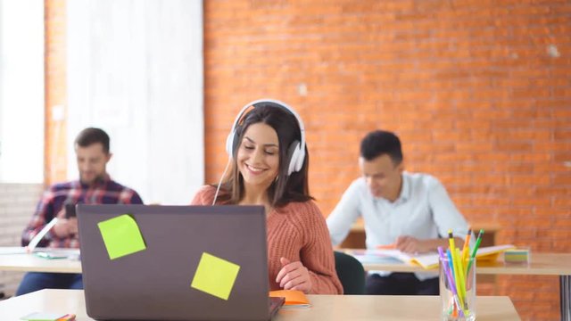 young happy brunette woman in big headphones sitting in front of laptop in office. Joyful girl dances and laughs at the desktop. In the background, young students of the mixed race learn. 4k
