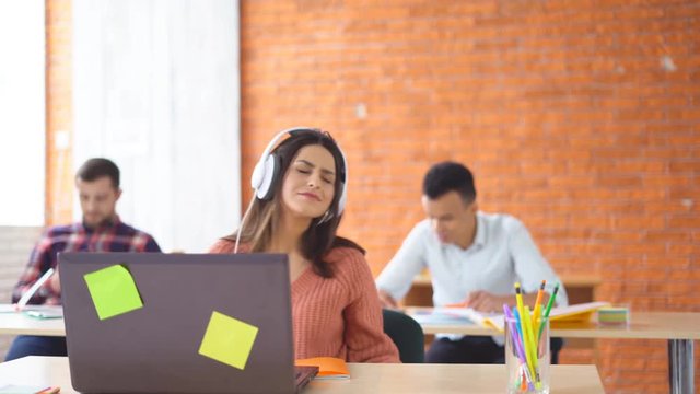 young happy brunette woman in big headphones sitting in front of laptop in office. Joyful girl dances and laughs at the desktop. In the background, young students of the mixed race learn. 4k